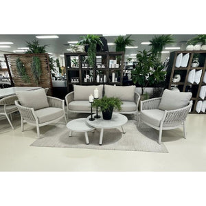 A 5-piece outdoor patio conversation set including a loveseat, two armchairs, and two nesting coffee tables, displayed in a showroom with other furniture and plants.